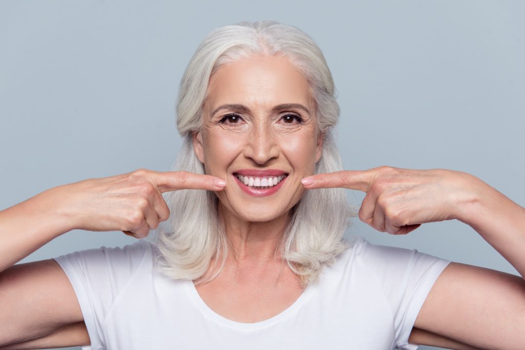 An older woman smiling and pointing to her white teeth with both hands