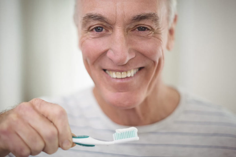 An older man smiles and holds a toothbrush with a glob of toothpaste on it