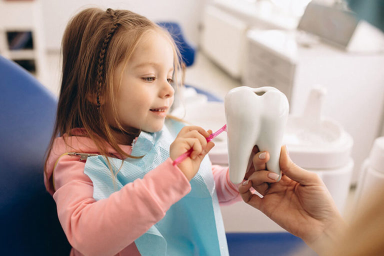 A pediatric dental patient sits in a dental exam chair and brushes a model of a tooth that an out of view adult is holding