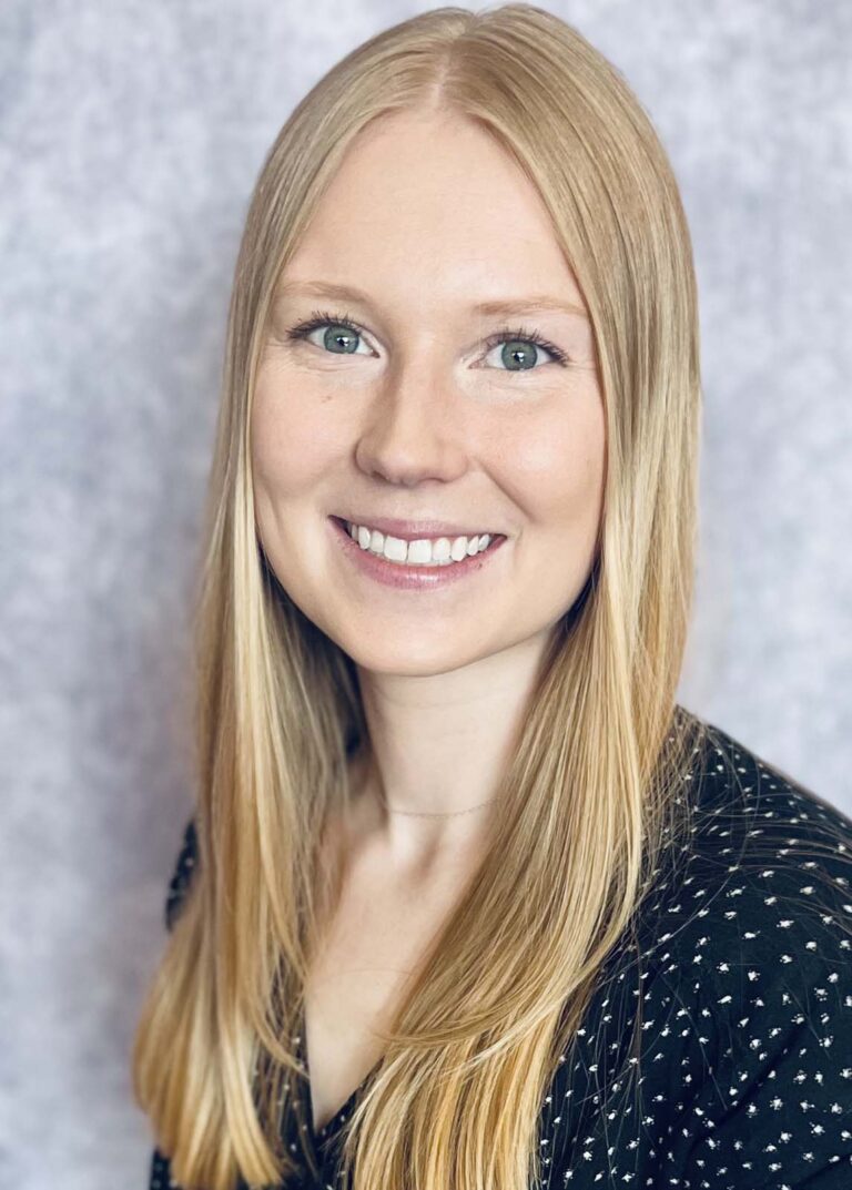 Portrait of Dr. Katie Sandefur, a dentist at Humlicek Dental, featuring her smiling broadly with light blonde hair, clear blue eyes, and wearing a dark blouse with a subtle floral pattern. The background is a soft, mottled grey.